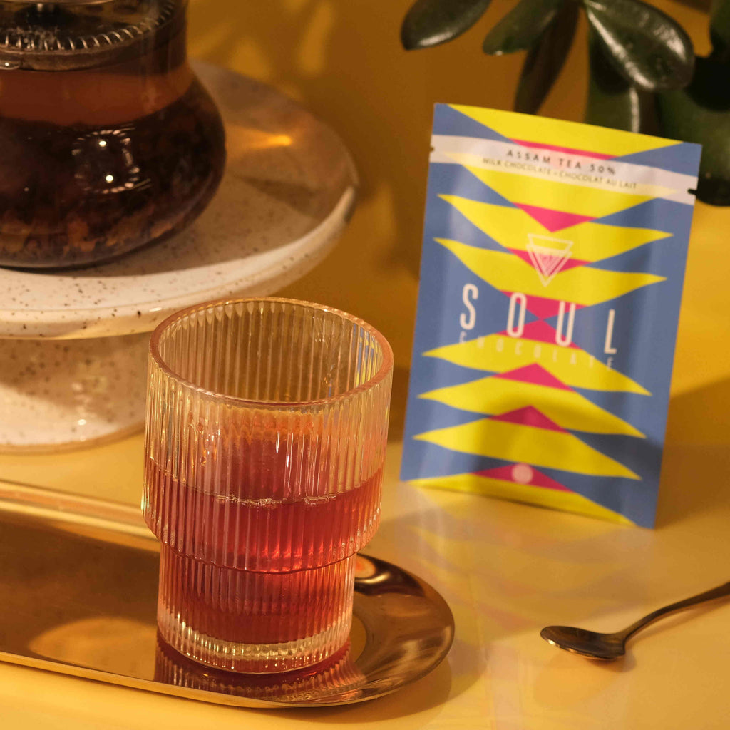 A glass cup of assam tea in a table setting near a wrapped bar of assam tea chocolate