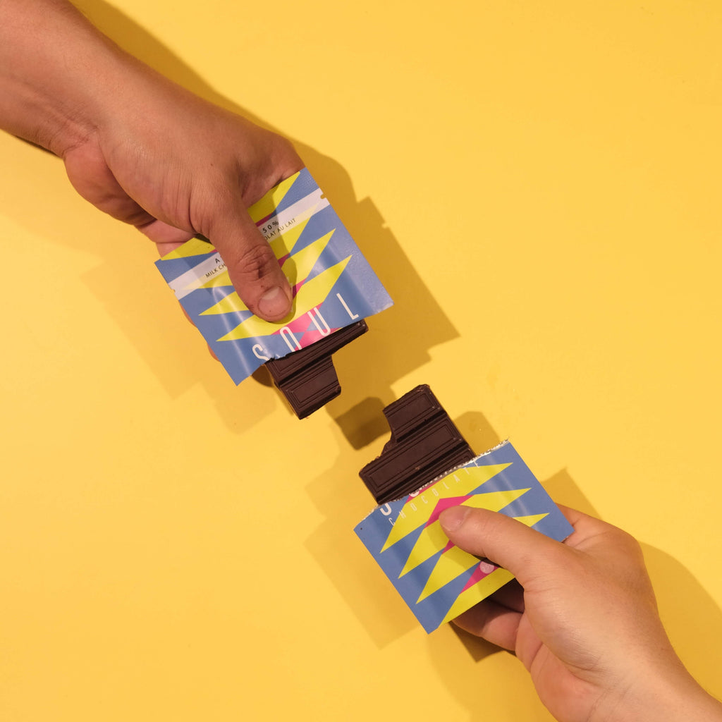 Two hands holding colorful unwrapped Assam chocolate bars against a yellow background