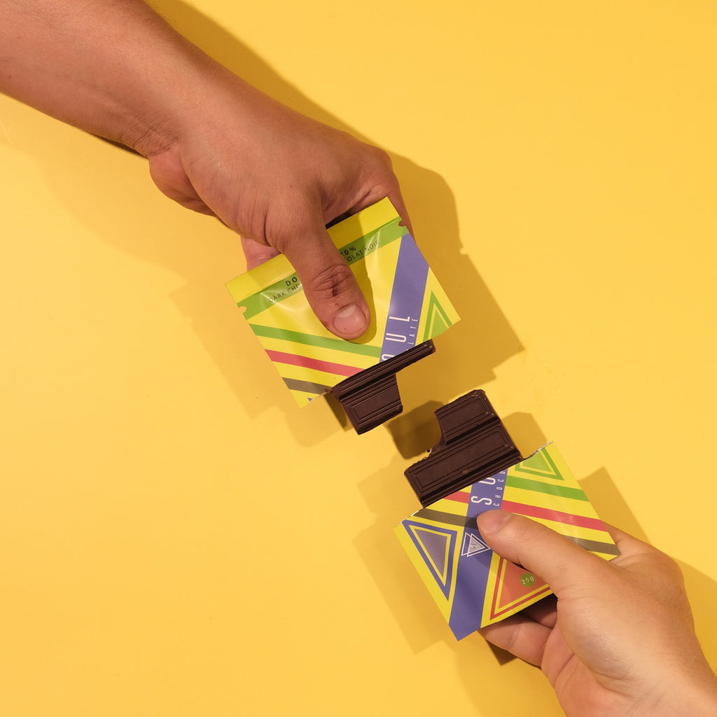 Two hands holding colorful unwrapped chocolates against a yellow background
