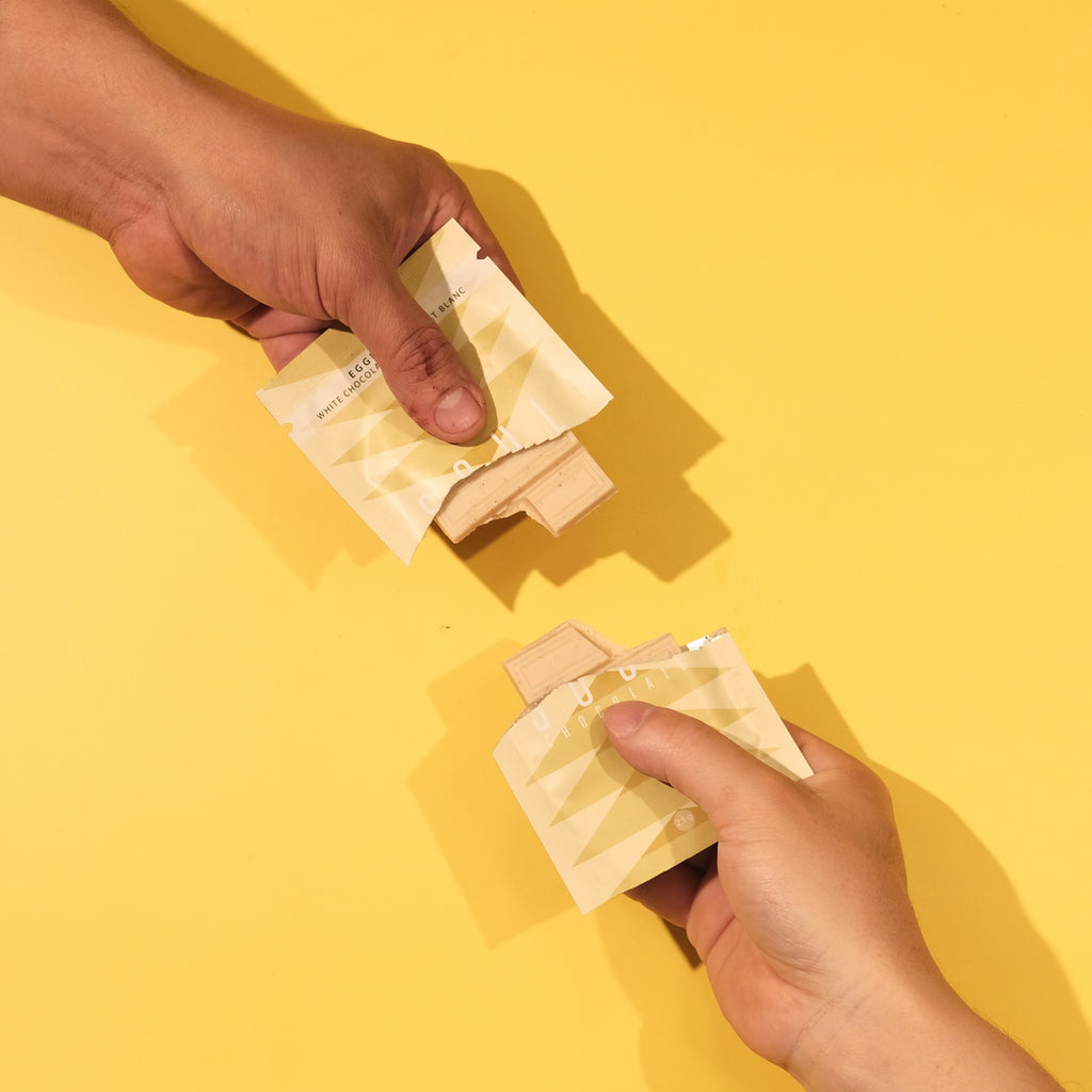 Two hands holding packets of eggnog chocolate against a yellow background