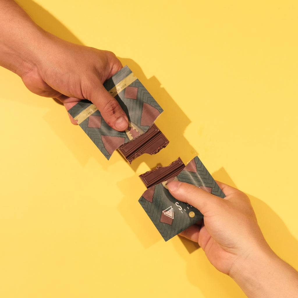 Two hands holding chocolate bars with decorative wrapping against a yellow background