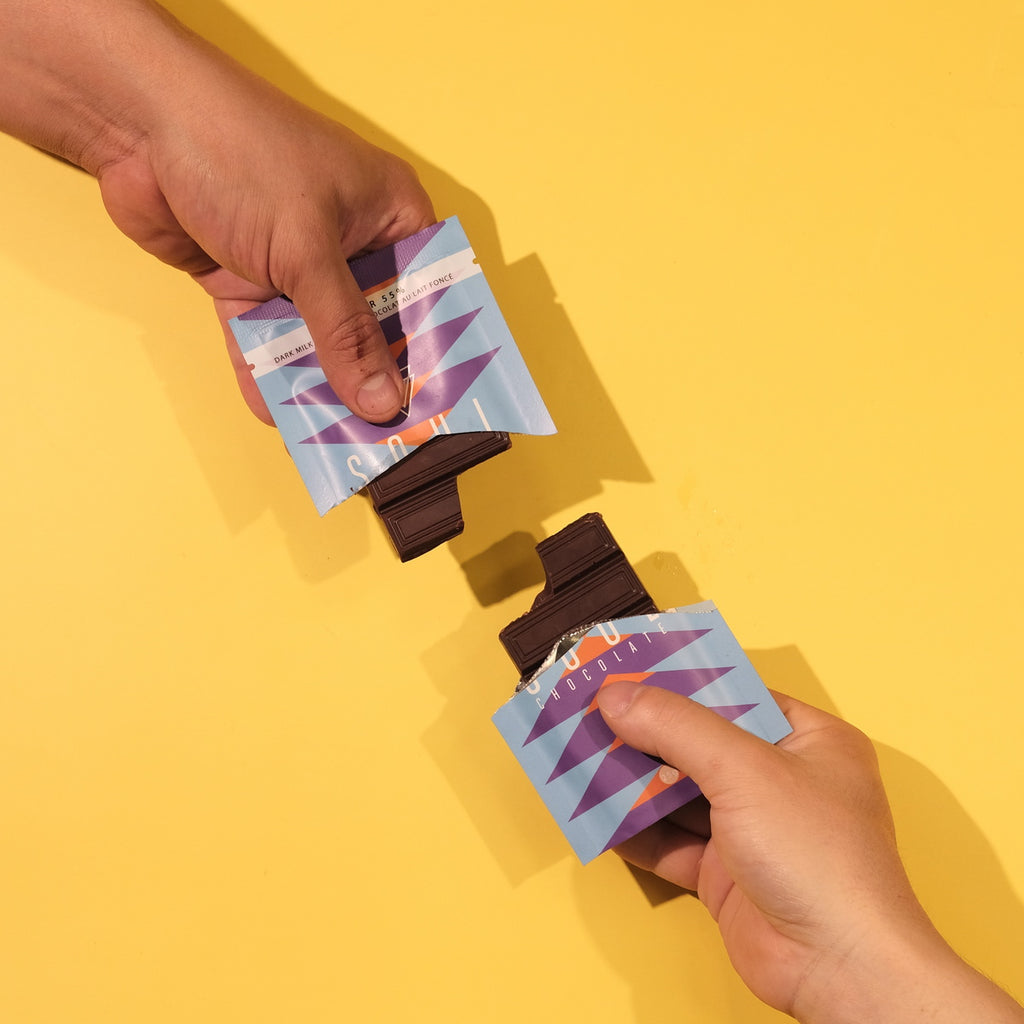 Two hands holding unwrapped Canadian Lavender chocolate with purple designs against a yellow background