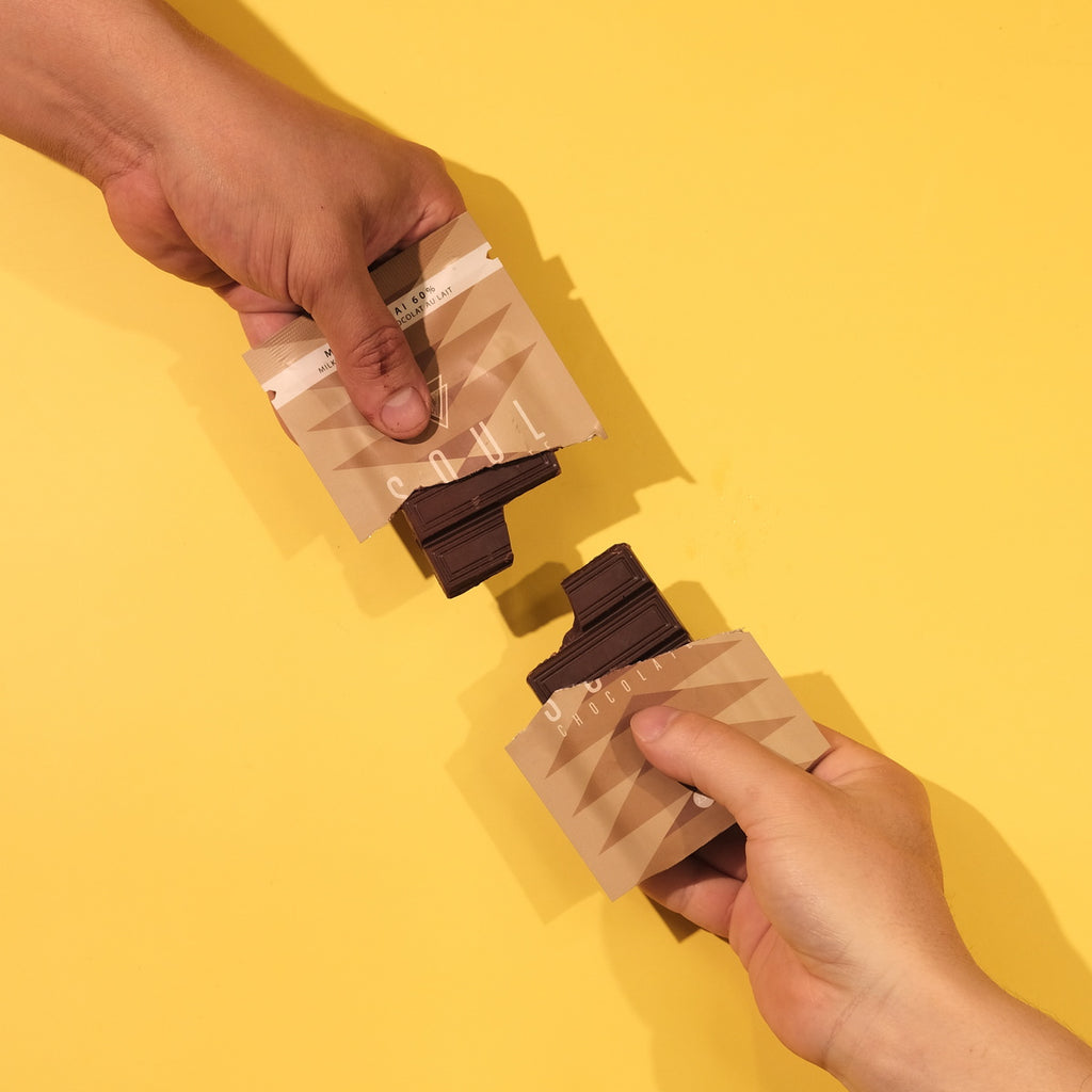 Two hands holding chai chocolate packets against a yellow background