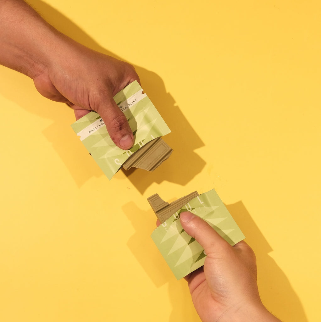 Two hands holding green packages of green tea matcha chocolate against a yellow background