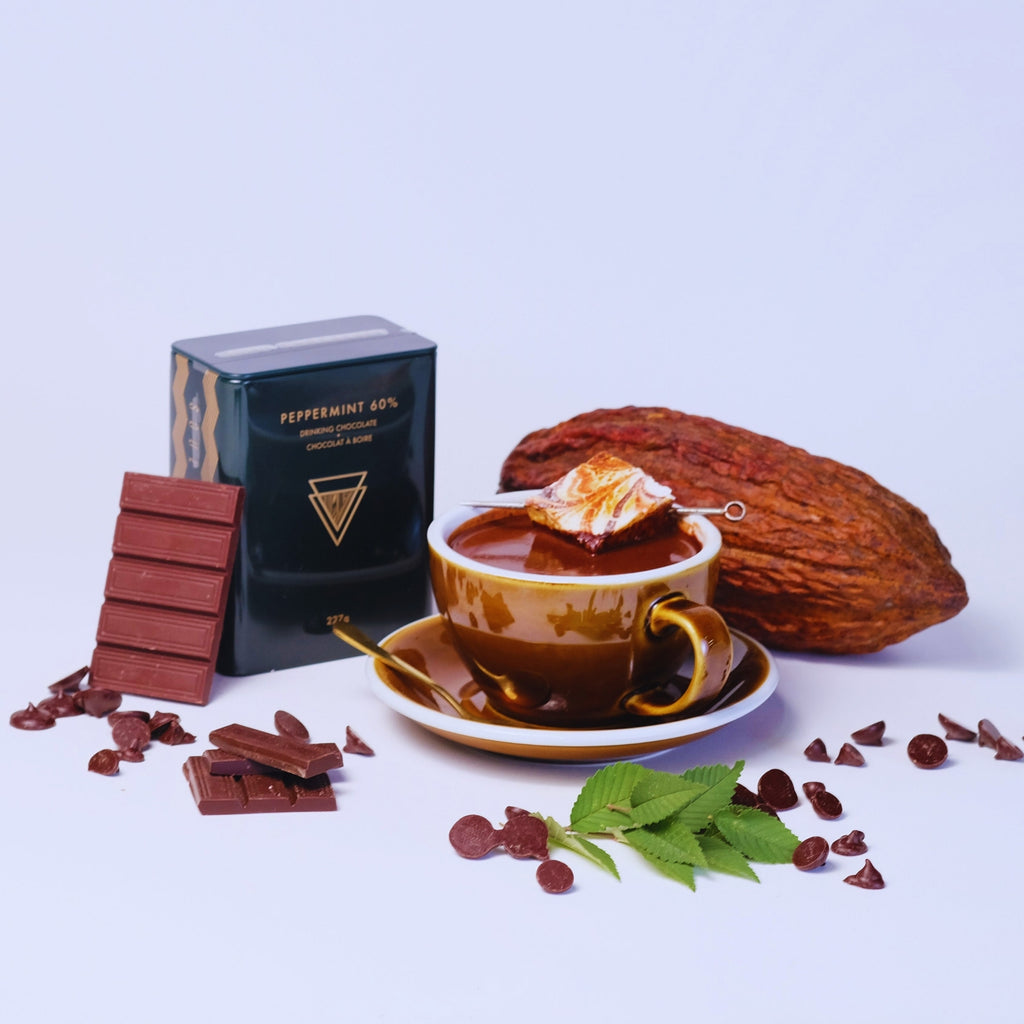 A warm mug of minty hot chocolate sits next to a cocoa pod and a tin of peppermint hot chocolate mix