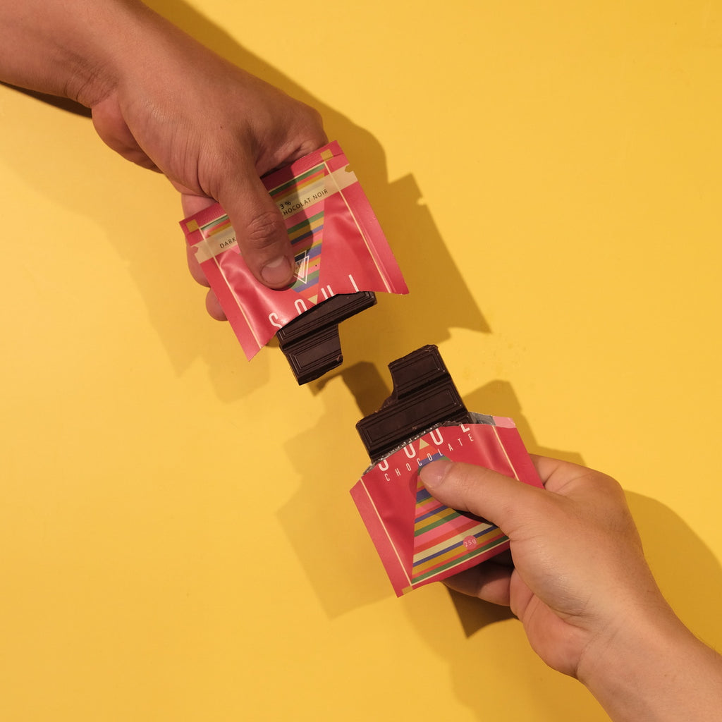 Two hands holding pink chocolate labeled packages with colorful labels against a yellow background