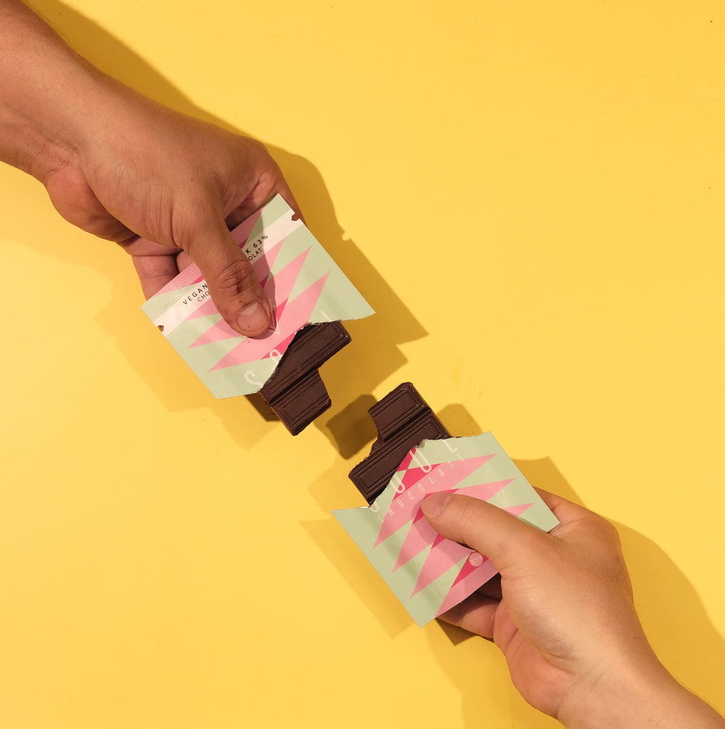 Two hands holding vegan unwrapped chocolate bars on a yellow background