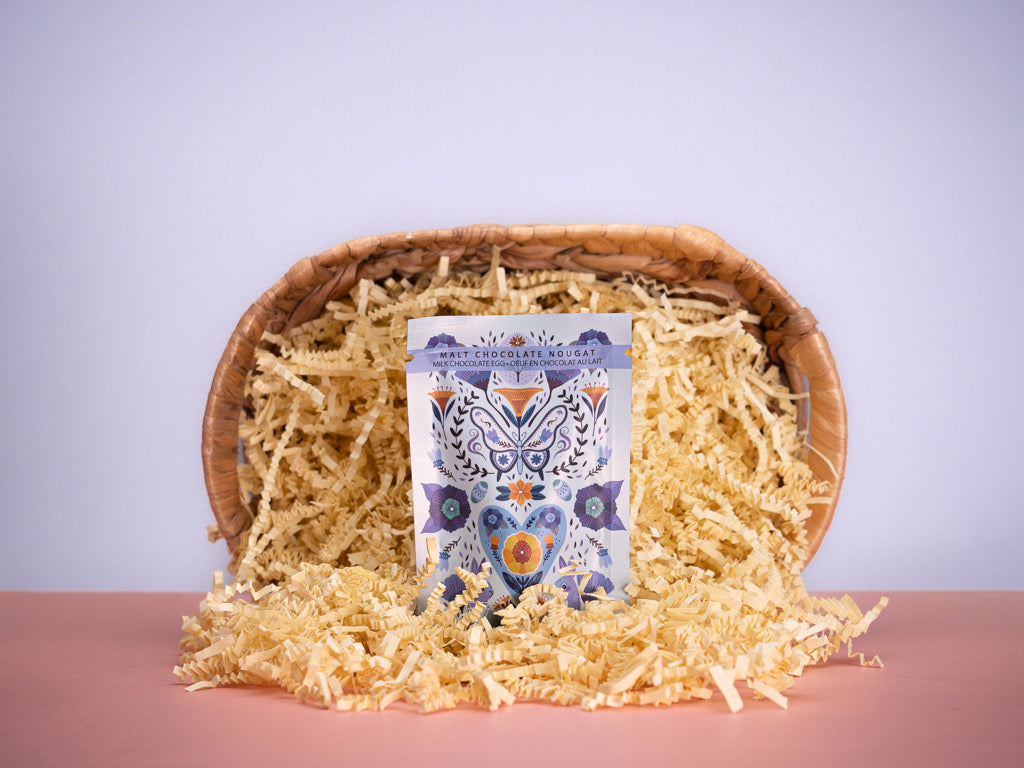 A chocolate egg with a patterned wrapper, placed on a bed of decorative straw within a wooden basket.