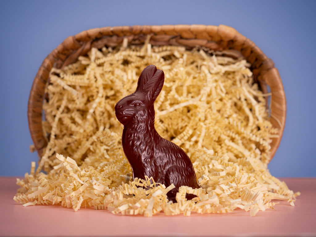 A solid chocolate Easter rabbit placed in a basket filled with straw, on a blue background.