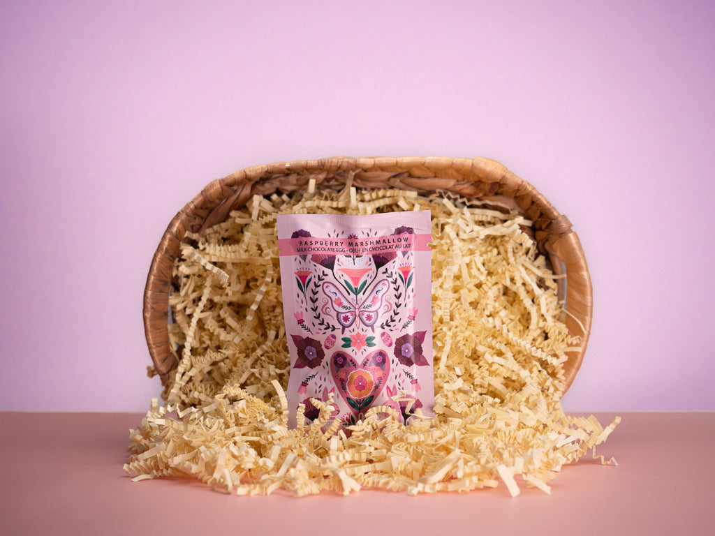 A chocolate egg with raspberry marshmallow, presented in a basket with decorative paper shreds, against a pink background.