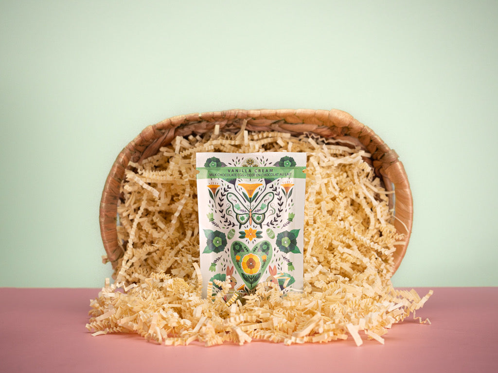 A chocolate egg with vanilla cream filling, salted caramel yolk, and a rich chocolate shell, decorated with colored cocoa butter, presented in a basket with decorative paper shreds.